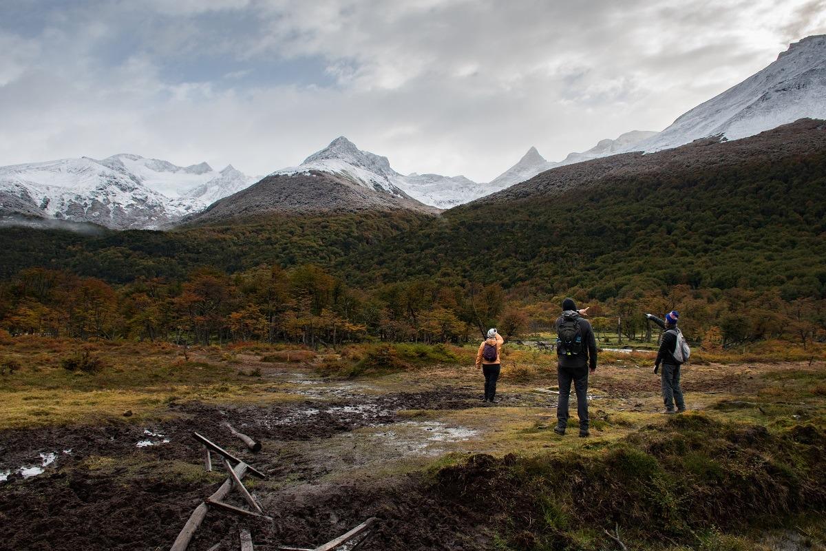 Three people on the muddy meadow, each showing to other direction. Snowy mountains in the distance