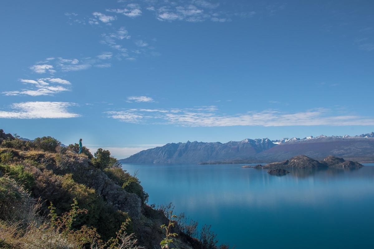 A blue lake in Chile, a girl in the distance, looking at it. A mountain range in the distance.